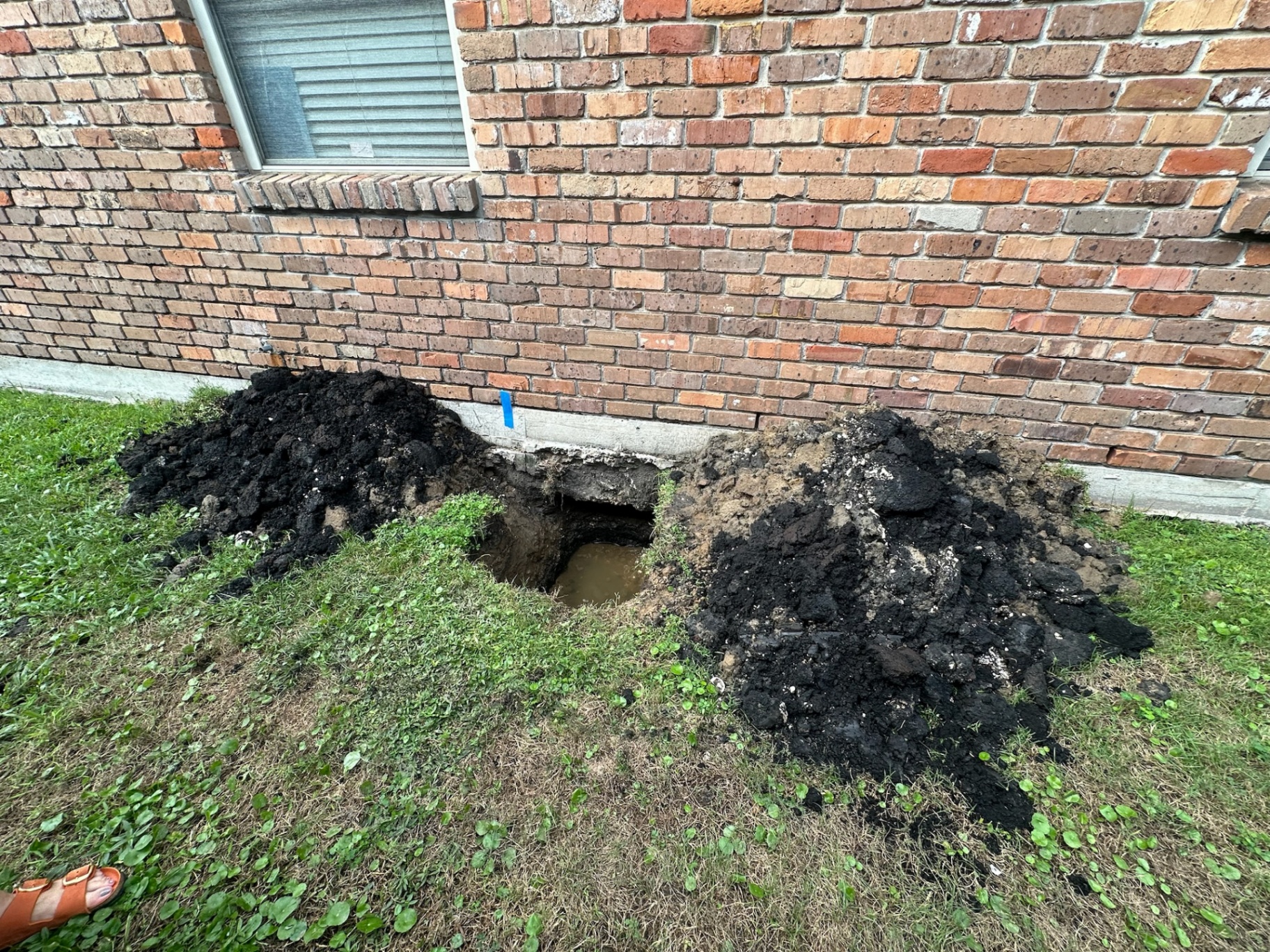 Hand-dug test pit at the base of the existing brick wall in Metairie with mounded spoil piles and standing groundwater in the bottom of the hole exposing how shallow the original footing sat before the second-floor addition foundation upgrades