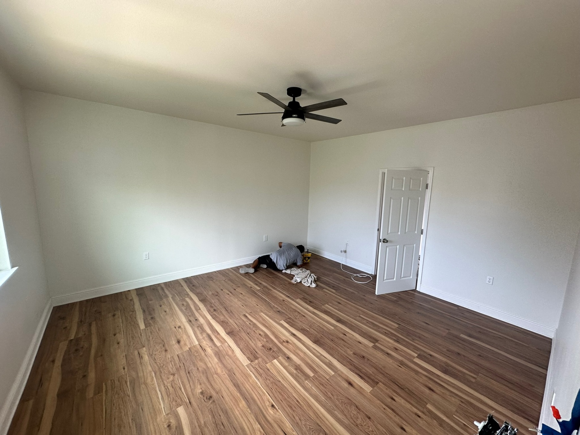 Finished new second-floor bedroom on the Metairie addition with newly installed wide-plank engineered wood flooring, white six-panel door, ceiling fan with light, fresh white walls and an installer transitioning trim along the baseboard