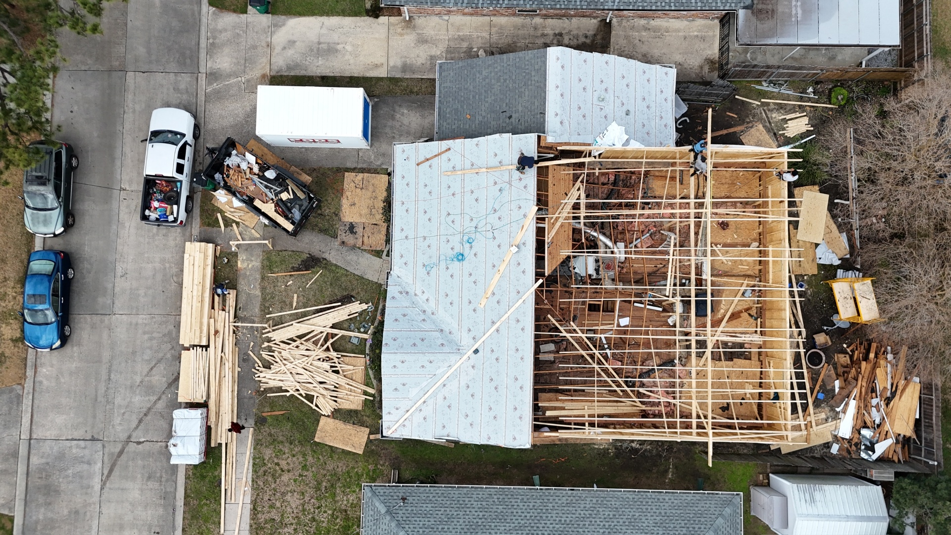Aerial drone view of Metairie home with PODS container and lumber delivery on the driveway as crew opens the roof and begins setting new second-floor framing