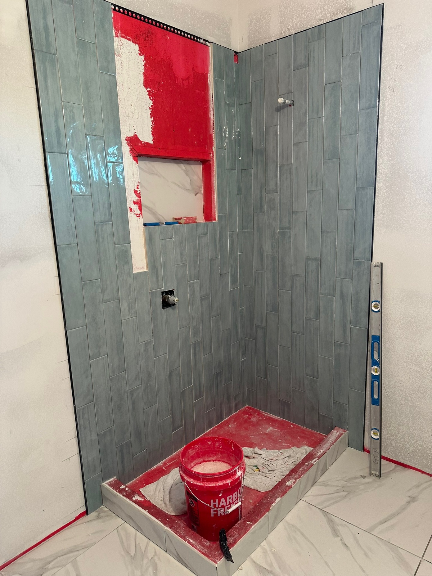 Master bathroom shower in the Metairie second-floor addition during tile installation with vertical sage-green glossy tile on two walls, RedGard waterproofing visible above the niche, marble shower pan and a level standing in the corner