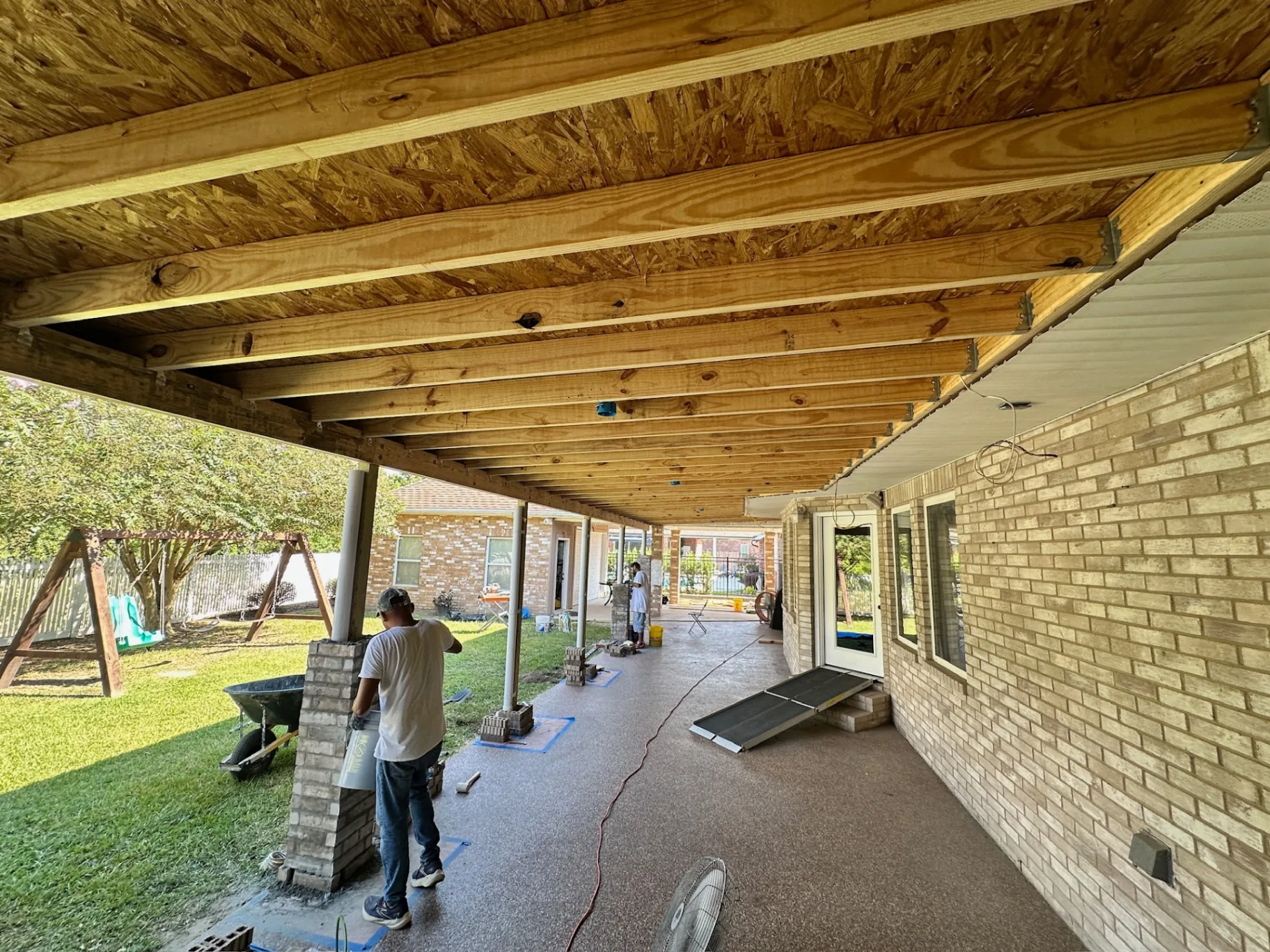 Underside view of new pressure-treated joists and decking with mason laying brick wrap around new patio posts at Destrehan Louisiana home