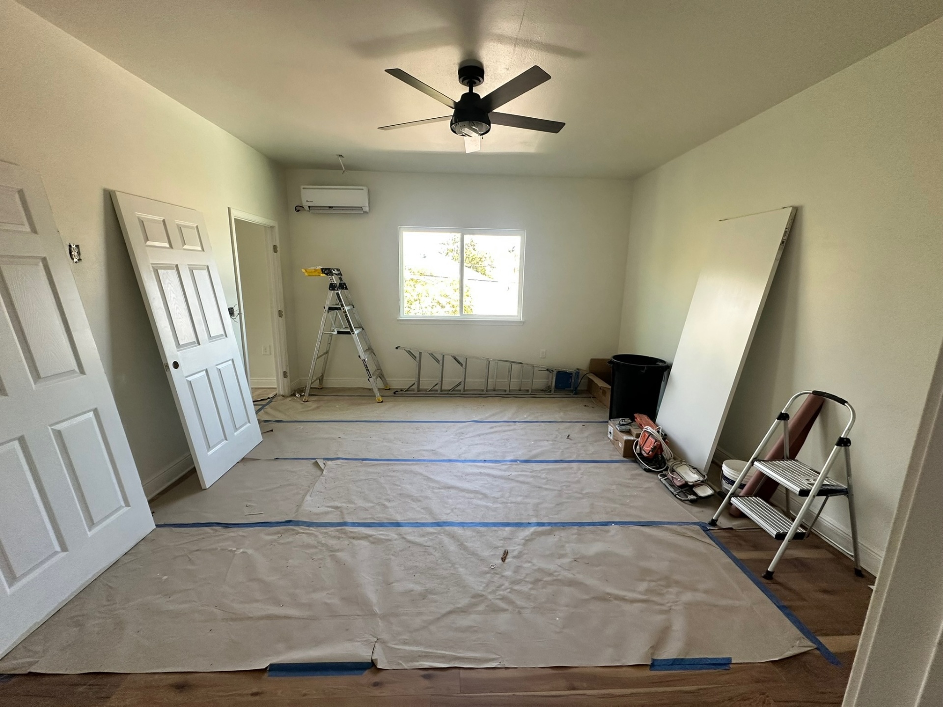 Finished second-floor bedroom with ductless mini-split head, ceiling fan, double-hung window, fresh paint and engineered hardwood floor with doors leaning ready to hang