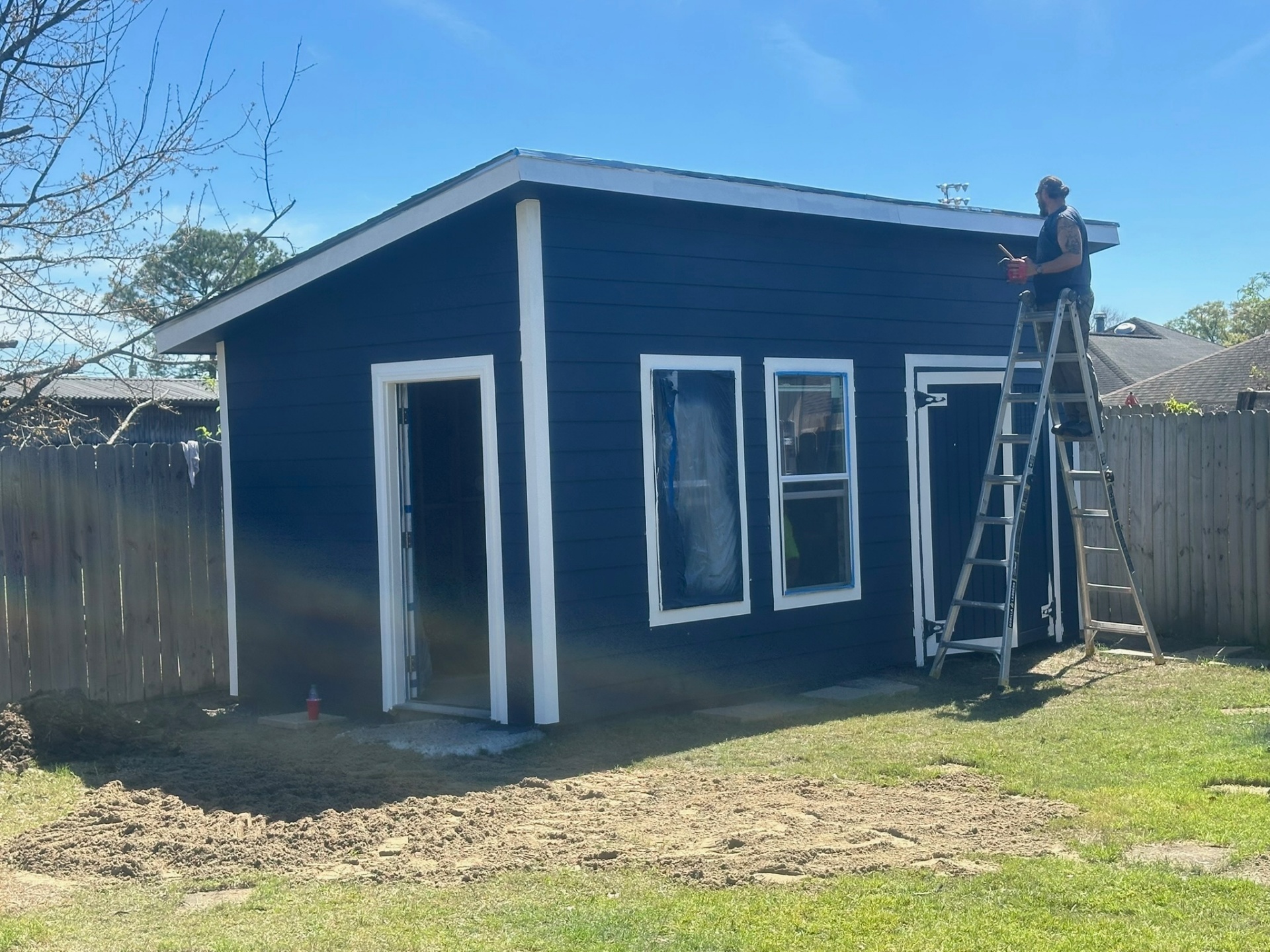 Cajun Home Services crew member on ladder painting fascia and drip edge on navy Hardie-siding shed in Metairie Louisiana