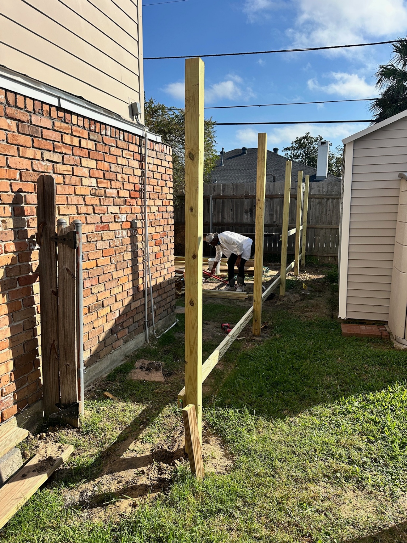 Freshly set pressure-treated 4x4 fence posts running along the side yard of the Bissonet Dr Metairie home during second-floor addition, with existing brick wall and shed visible