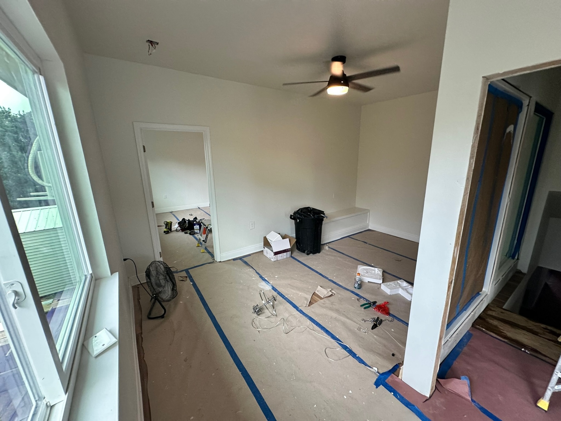 New second-floor bedroom on the Metairie addition during finish stage with paper floor protection taped down, ceiling fan, fresh white walls, large window over the back of the house and a doorway into a second new bedroom beyond