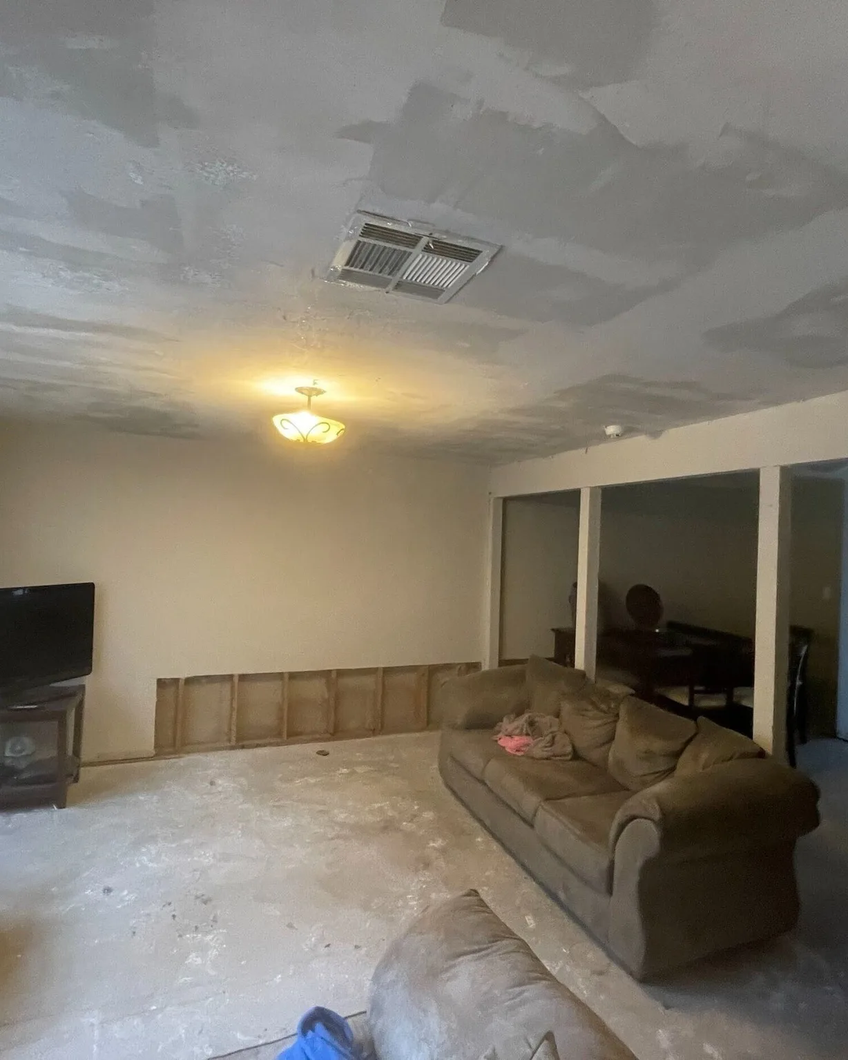 Gretna LA living room before remodel showing water-damaged ceiling with patched sheetrock, removed baseboard drywall from flood damage, dusty concrete floor and old couch — Cajun Home Services water damage and interior remodel project in Jefferson Parish