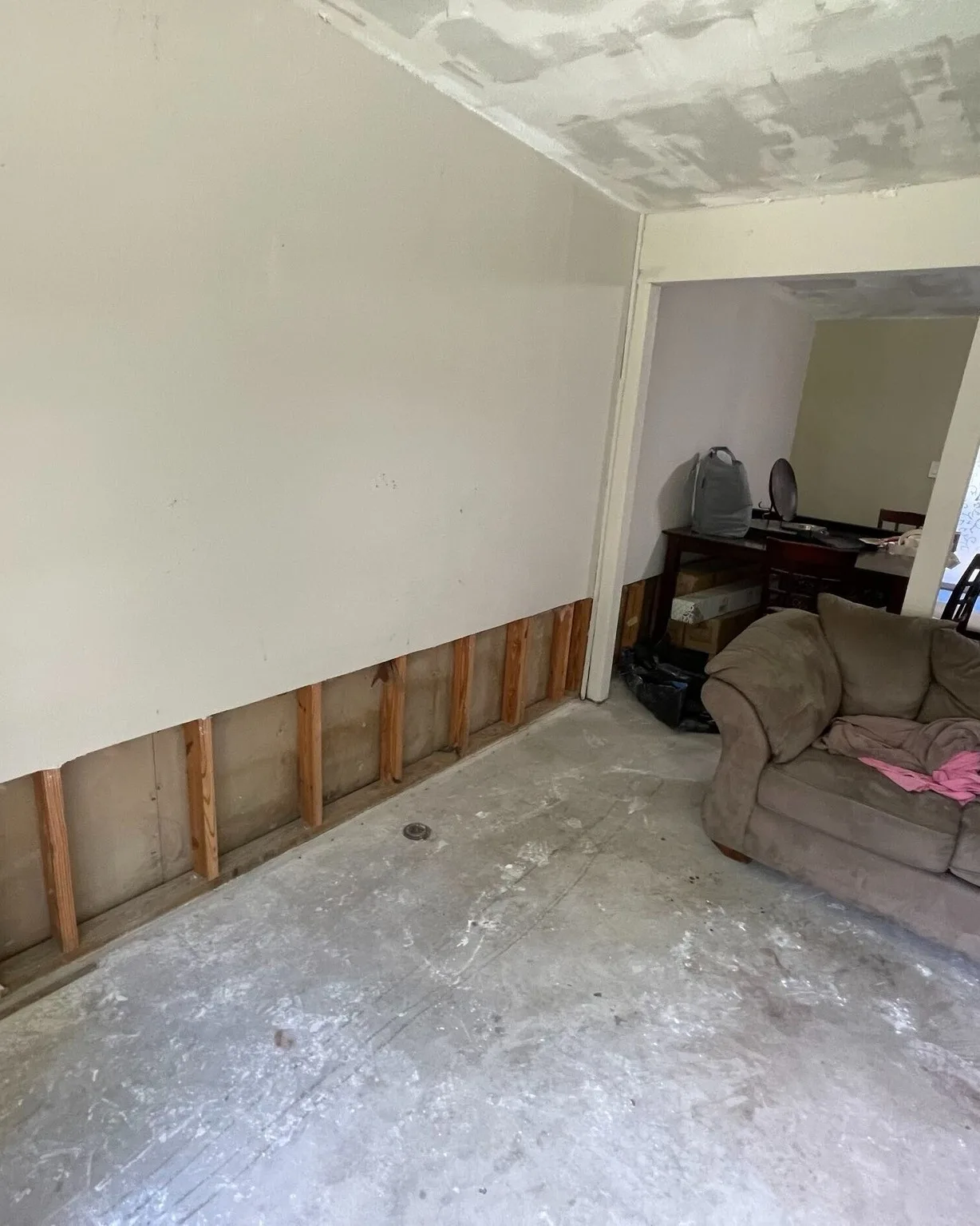 Gretna LA living room before remodel showing exposed wall framing at base where flood-damaged sheetrock was cut out, bare concrete slab and unfinished ceiling during demo phase by Cajun Home Services