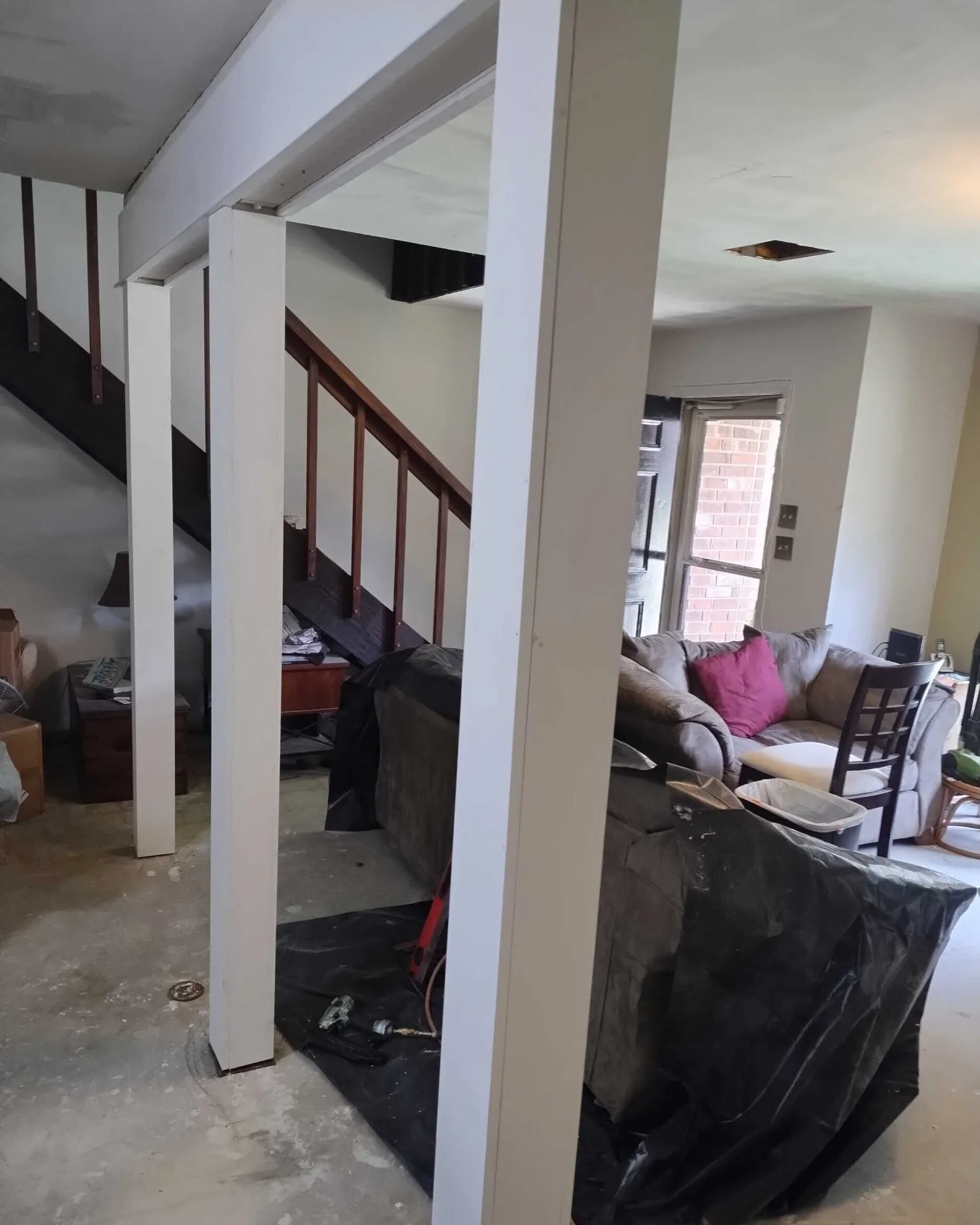 Gretna LA living room during demo with original wooden stair balustrade visible, raw structural columns wrapped in primed white casing and plastic-covered furniture protecting belongings during Cajun Home Services interior remodel