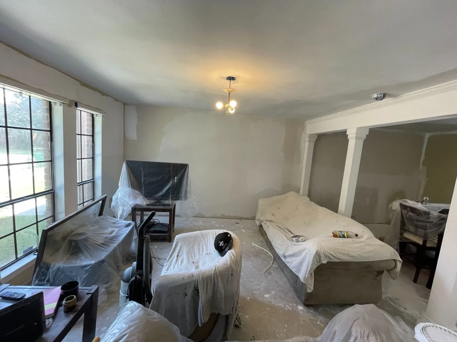 Gretna LA living room with skim-coated walls and ceiling fully prepped for paint, furniture protected by drop cloths and plastic sheeting, custom column trim visible at right by Cajun Home Services