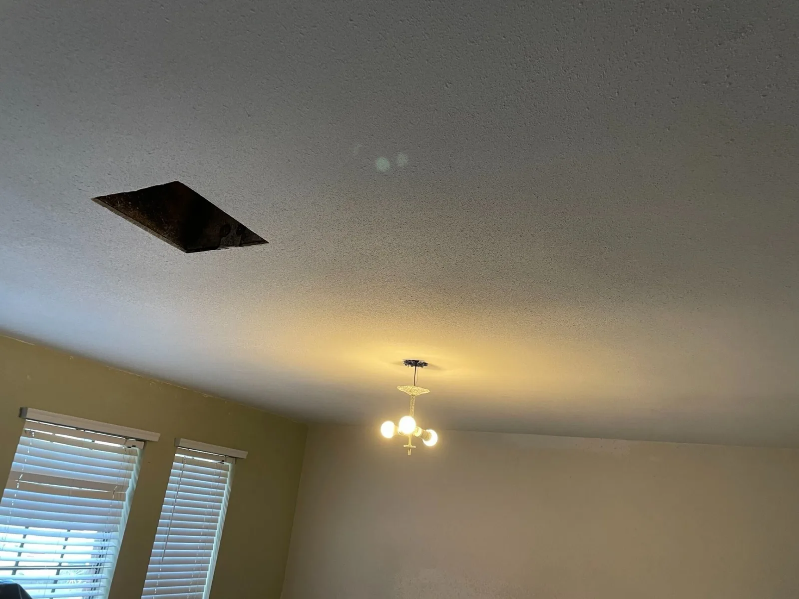 Gretna LA living room ceiling with cut-out access hole exposing attic framing next to old brass chandelier — investigating concealed water damage and wiring before patch and repaint by Cajun Home Services