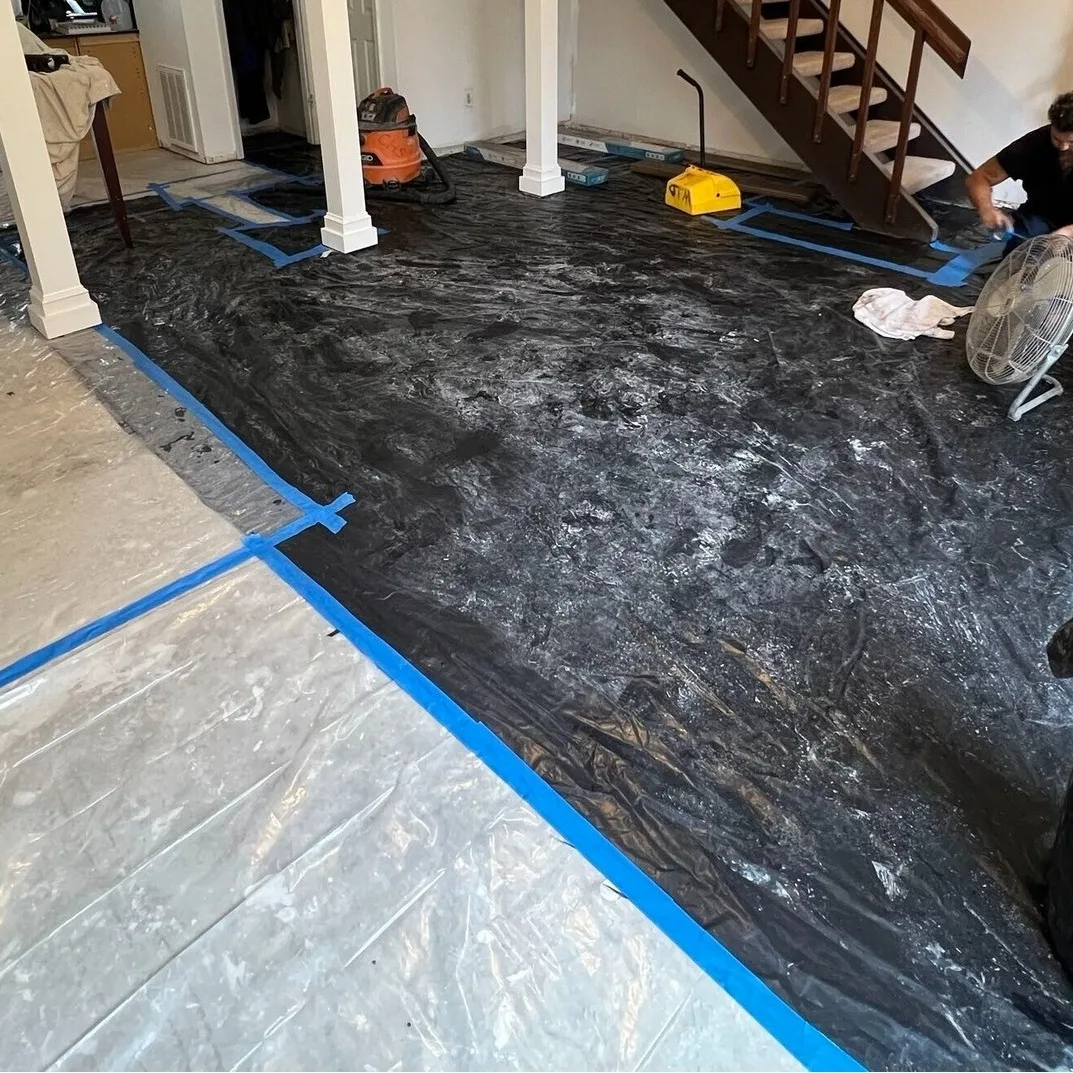 Gretna LA living room with black 6-mil moisture barrier rolled out over concrete slab, seams taped with blue painter tape, crew member operating air mover near staircase before LVP plank installation by Cajun Home Services