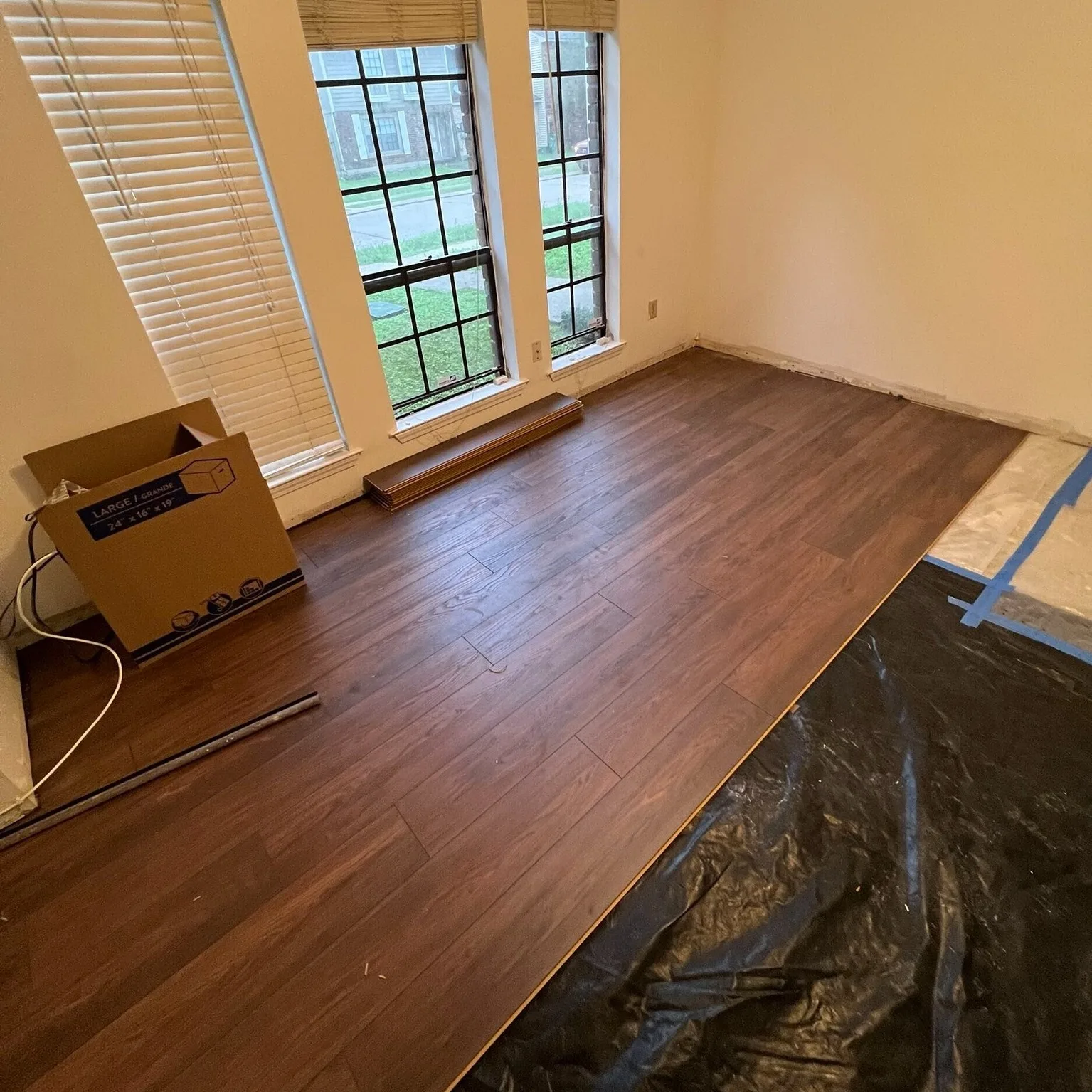 Gretna LA bay-window front room with newly installed warm walnut luxury vinyl plank flooring laid in straight pattern, stacked planks staged for next row, moisture barrier visible at right by Cajun Home Services
