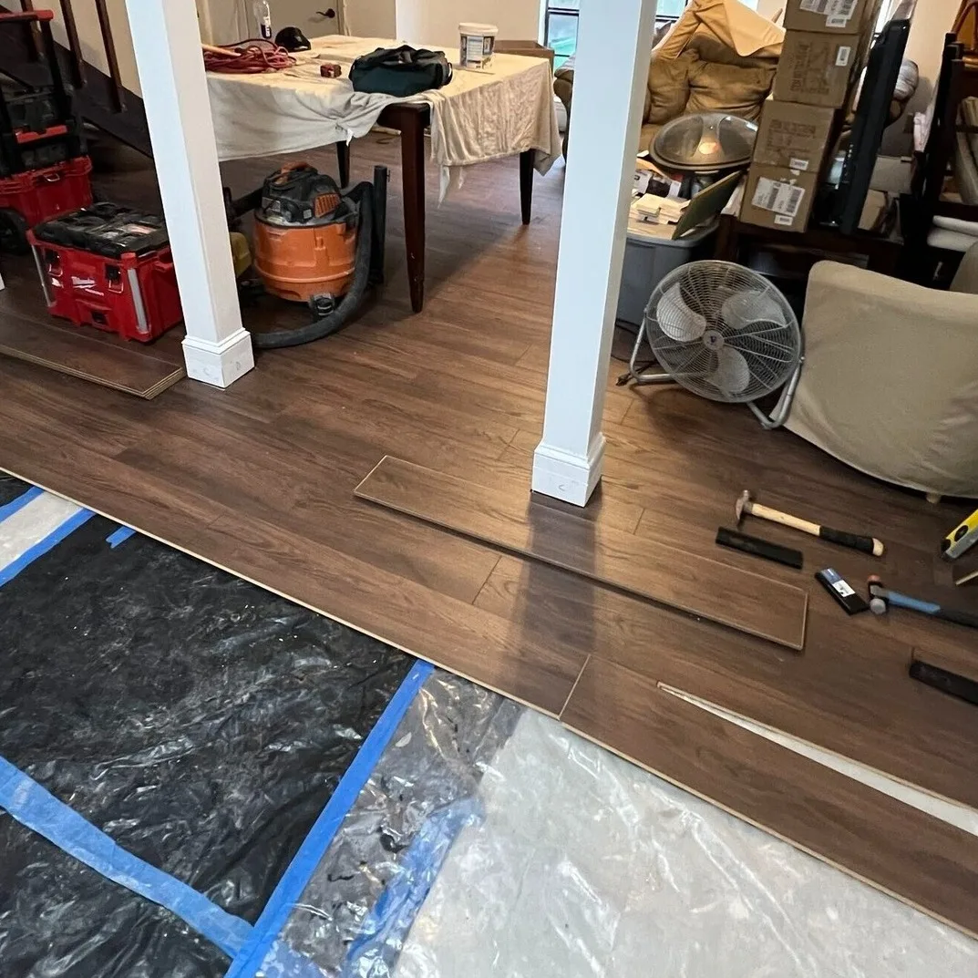 Gretna LA open living and dining room mid-LVP install with rich walnut planks running through column-framed openings, Milwaukee Packout tools and tapping block staged on remaining moisture barrier by Cajun Home Services