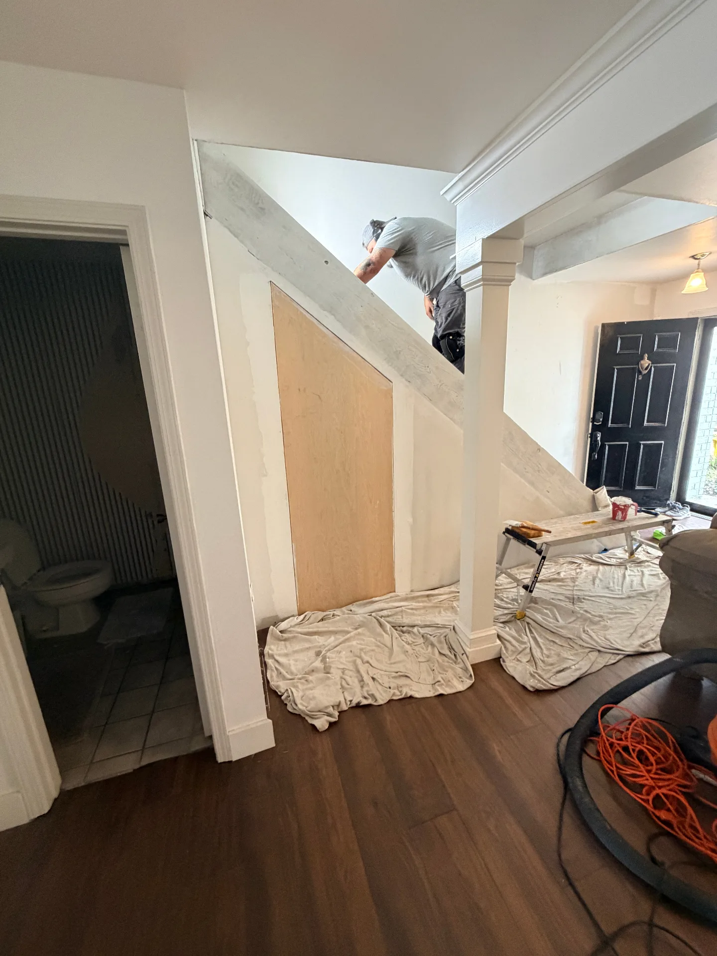 Gretna LA entryway under the staircase with Cajun Home Services carpenter installing custom plywood wainscoting panel along the angled stringer, drop cloths protecting new LVP floor by the front door