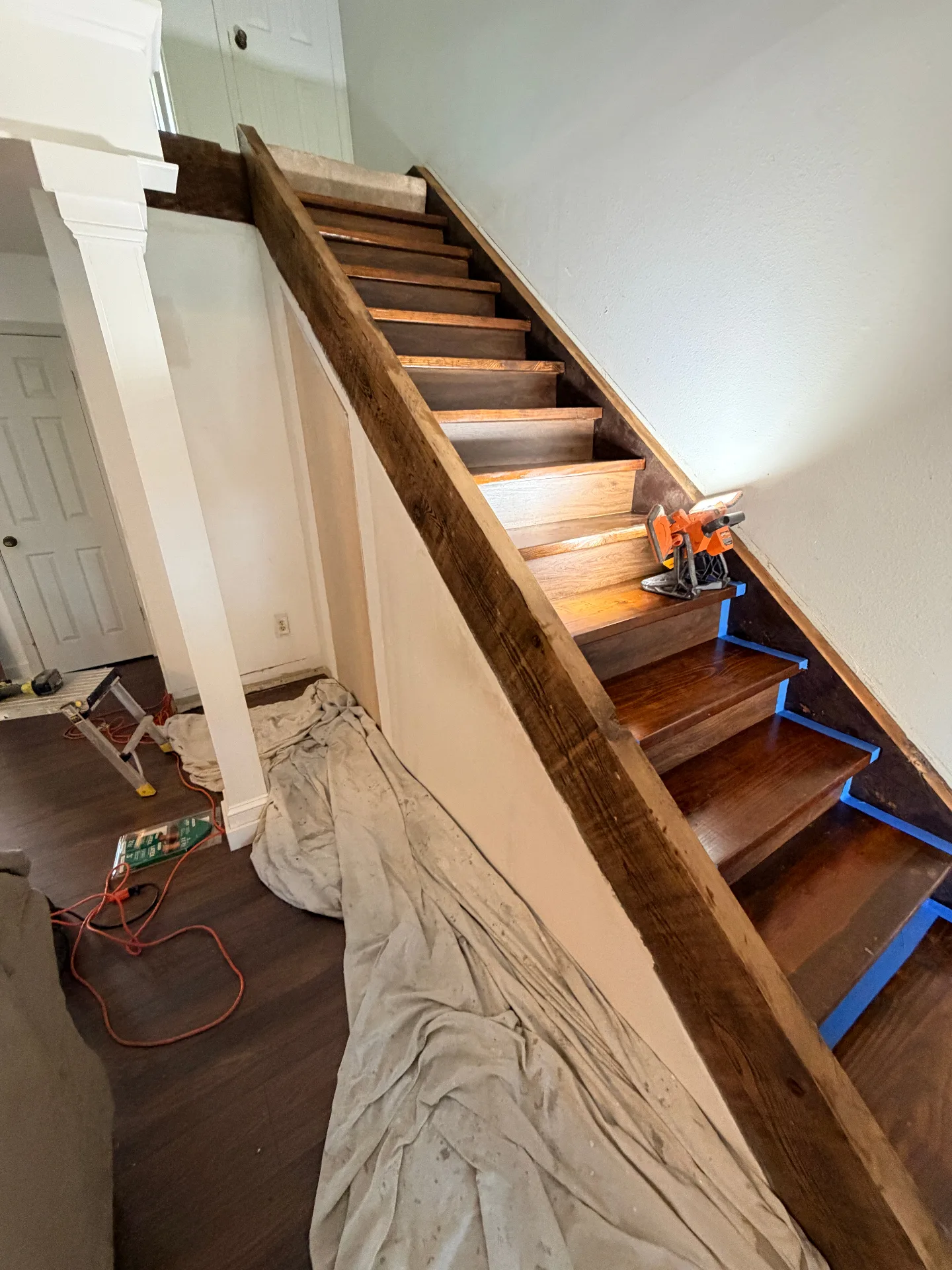 Gretna LA original wooden staircase mid-refinish with treads sanded back to raw oak and risers taped off in blue painter tape, deep walnut stain being applied step by step by Cajun Home Services