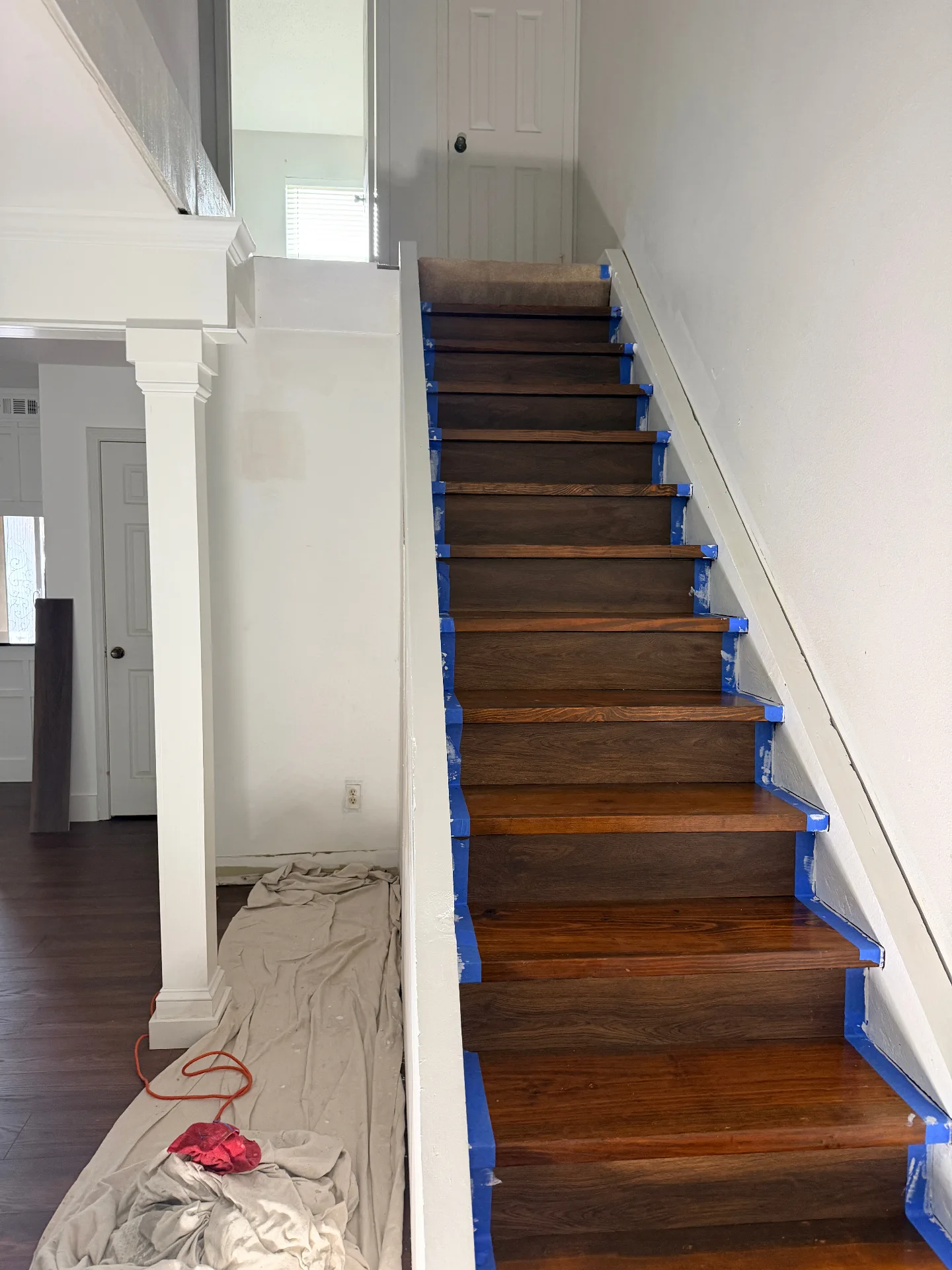 Gretna LA finished refinished oak staircase with rich glossy walnut treads and crisp white risers protected by blue tape, fresh trim and column wraps flanking the base — Cajun Home Services carpentry detail