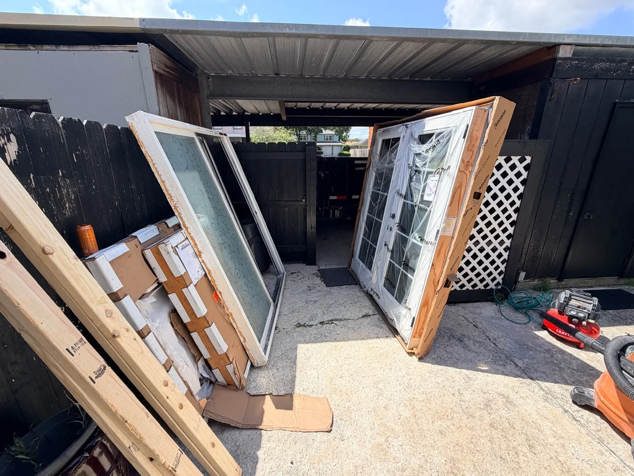 Gretna LA carport staging area with old aluminum patio slider removed and new ProVia French doors and replacement windows wrapped in factory packaging, ready for installation by Cajun Home Services