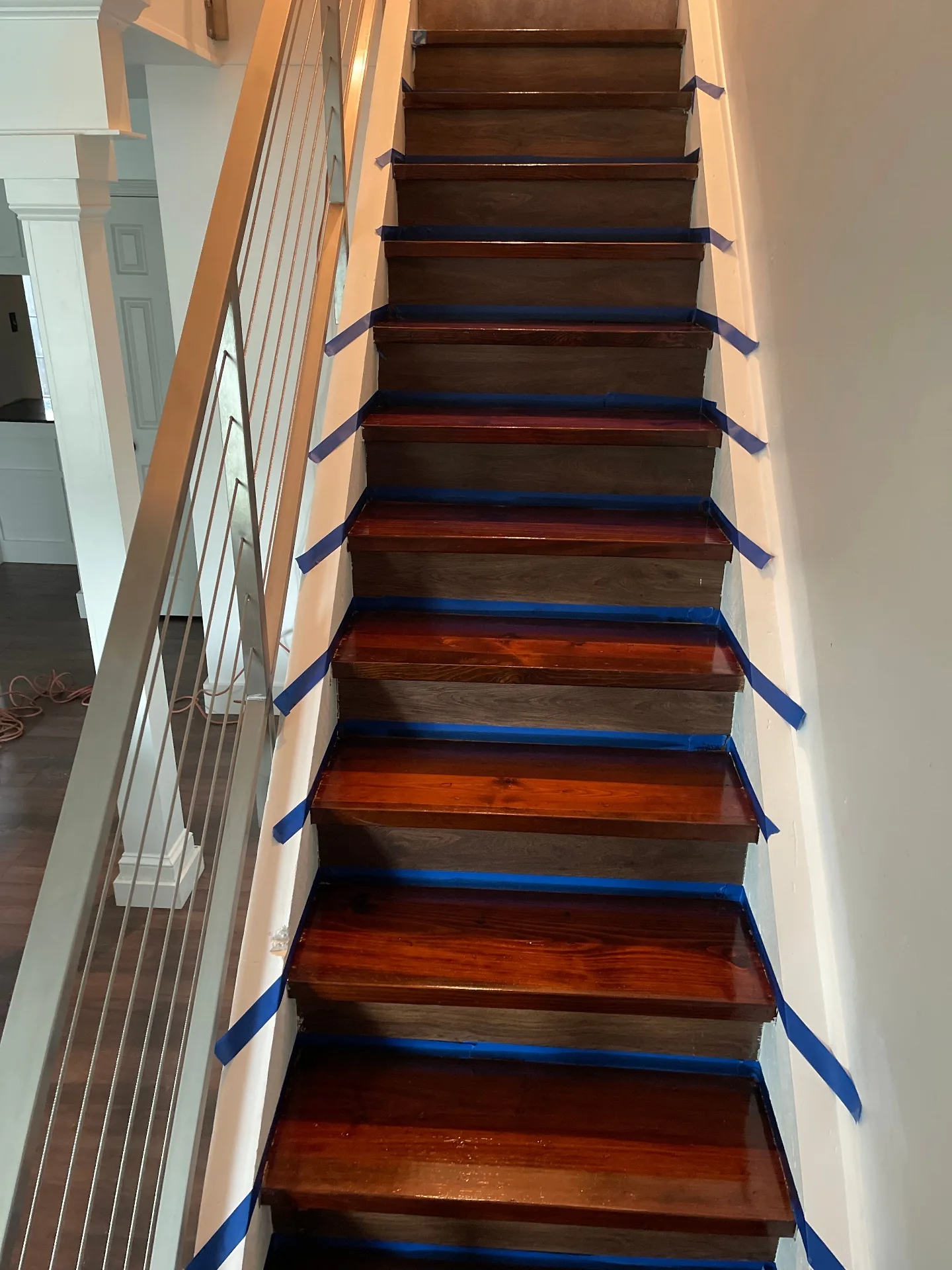 Gretna LA staircase mid-refinish showing transition from original glossy red-stained pine treads at the bottom to freshly applied dark walnut stain at the top, blue painter tape protecting white risers by Cajun Home Services