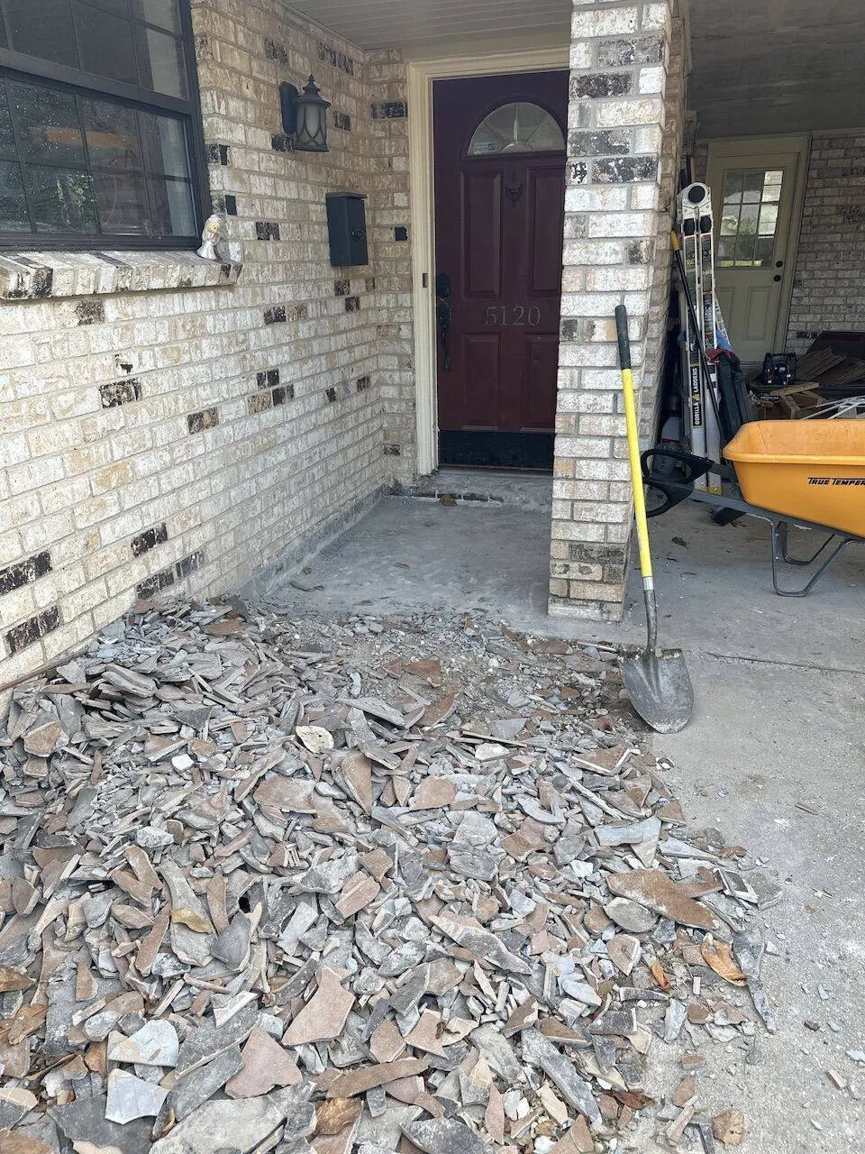 Front porch tile demolition with broken tile debris wheelbarrow and tools at brick home in Metairie