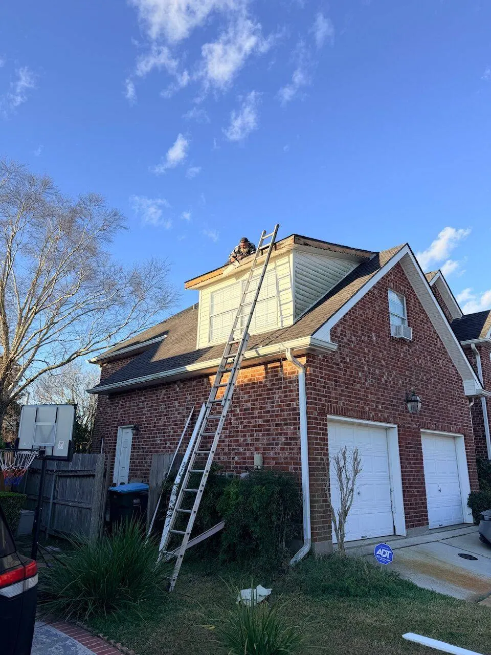 Crew member on roof of two-story brick home during shingle replacement with ladders in place