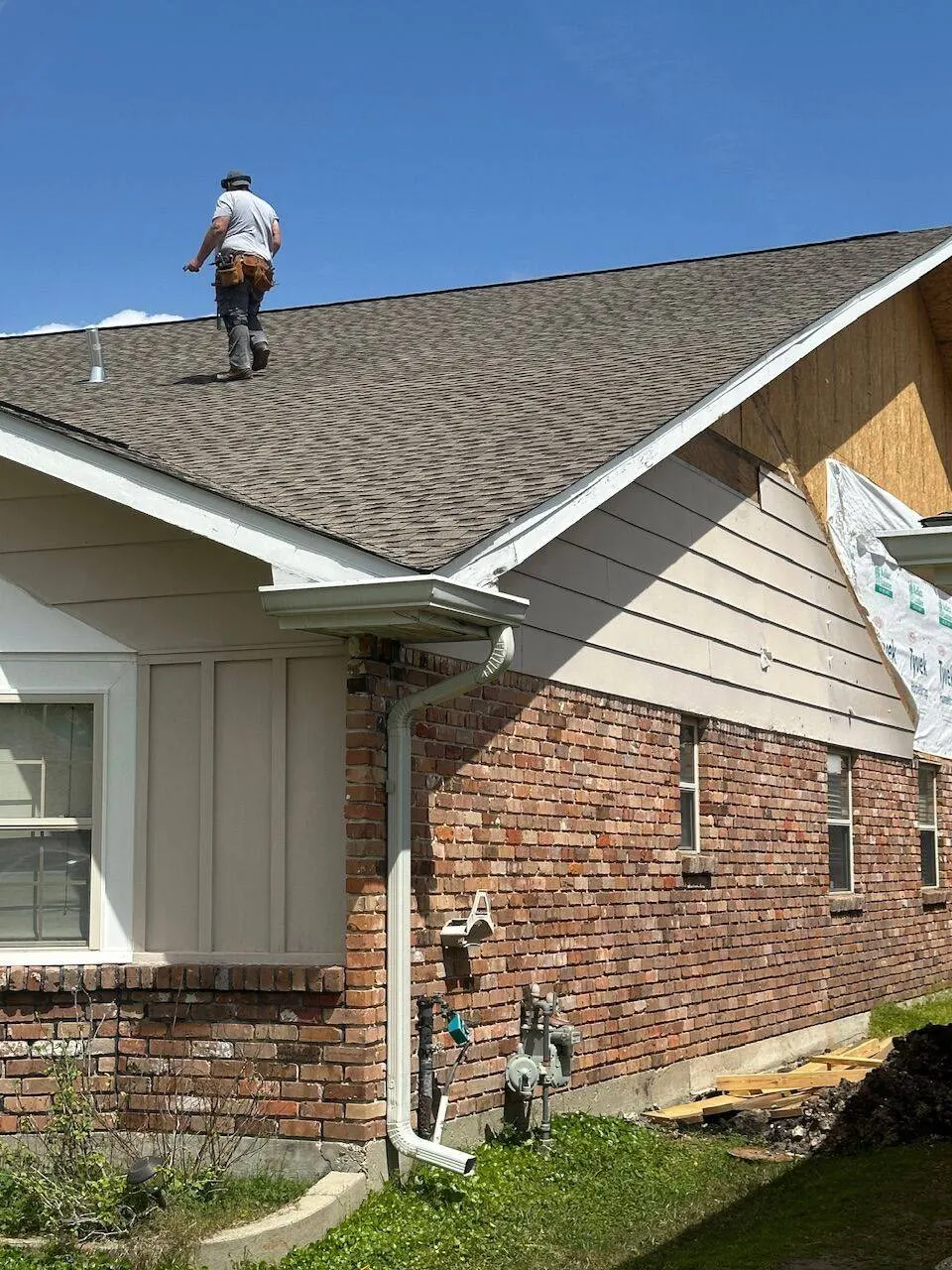 Roofer walking ridge of brick home during shingle replacement with tool belt and Tyvek wrap visible