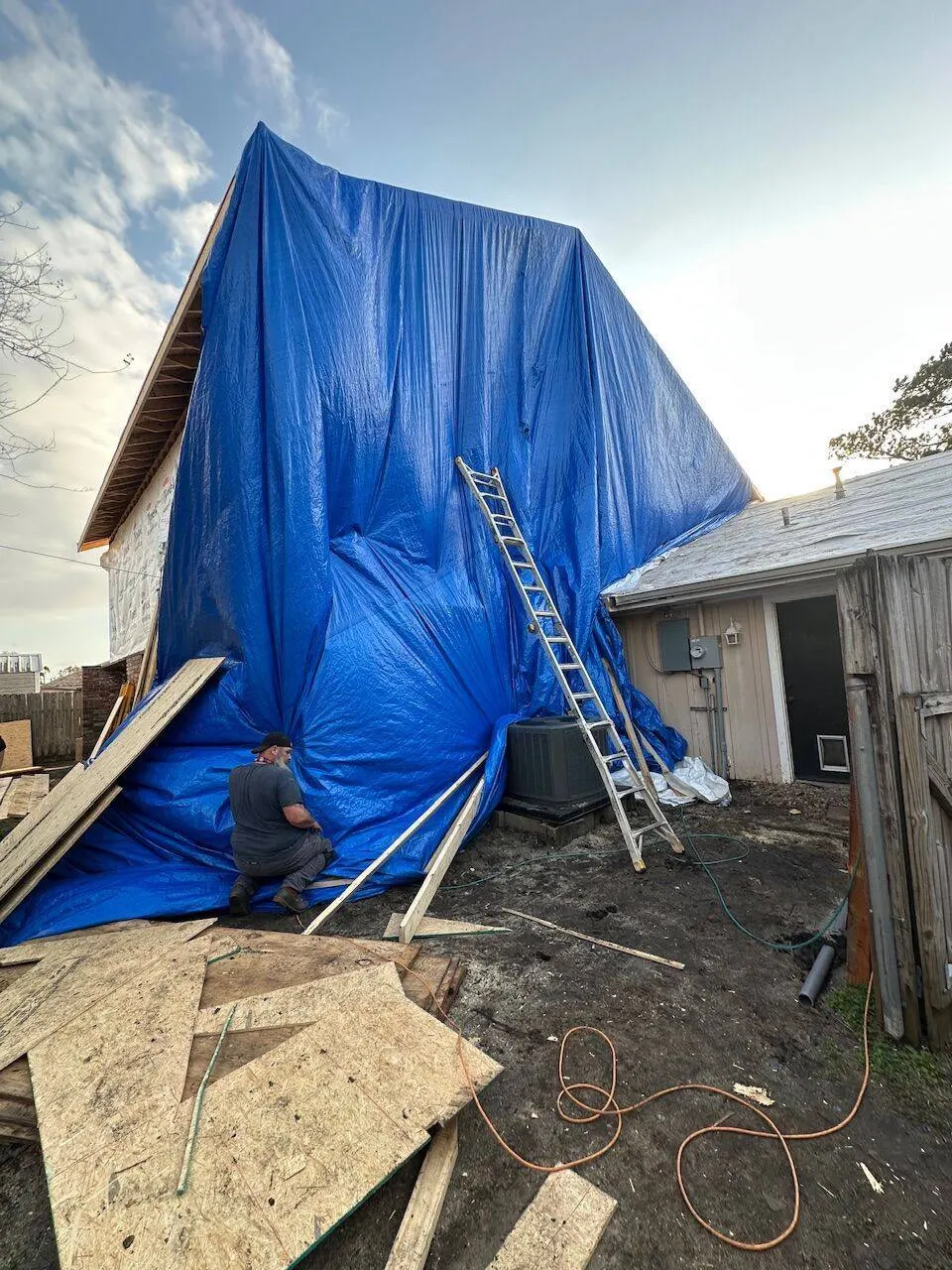 Blue tarp protecting home during active reroof with ladder and crew worker securing protection
