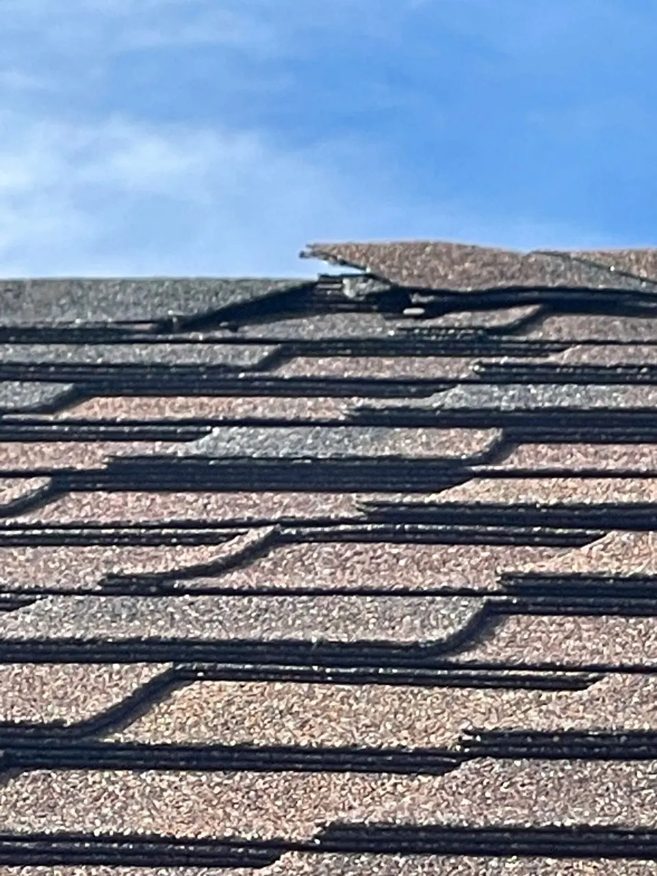Close-up of cracked and broken ridge cap shingle tab on aged Destrehan Louisiana asphalt shingle roof, exposing the asphalt mat beneath the granule surface before full re-roof replacement