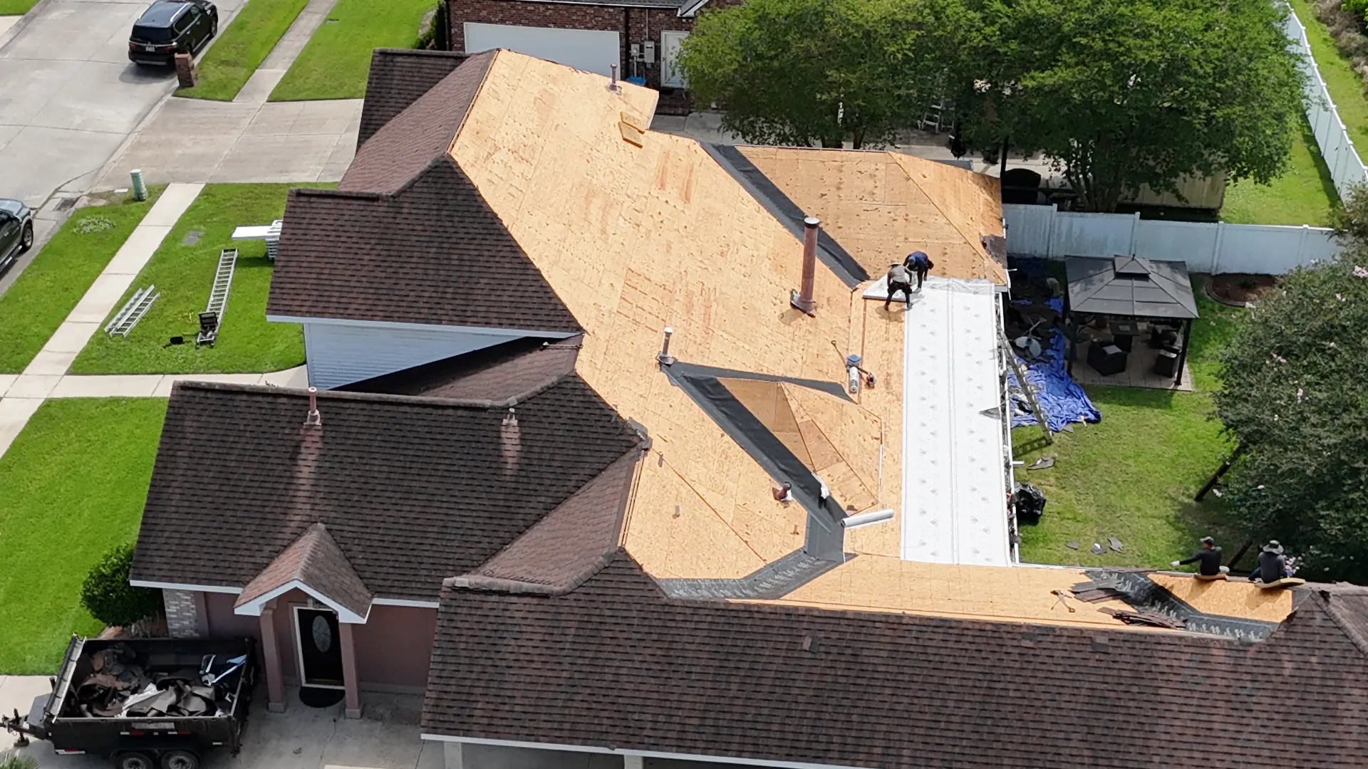 Aerial view of exposed OSB roof decking after shingle tear-off on a Destrehan re-roofing project, with debris dump trailer parked at the driveway