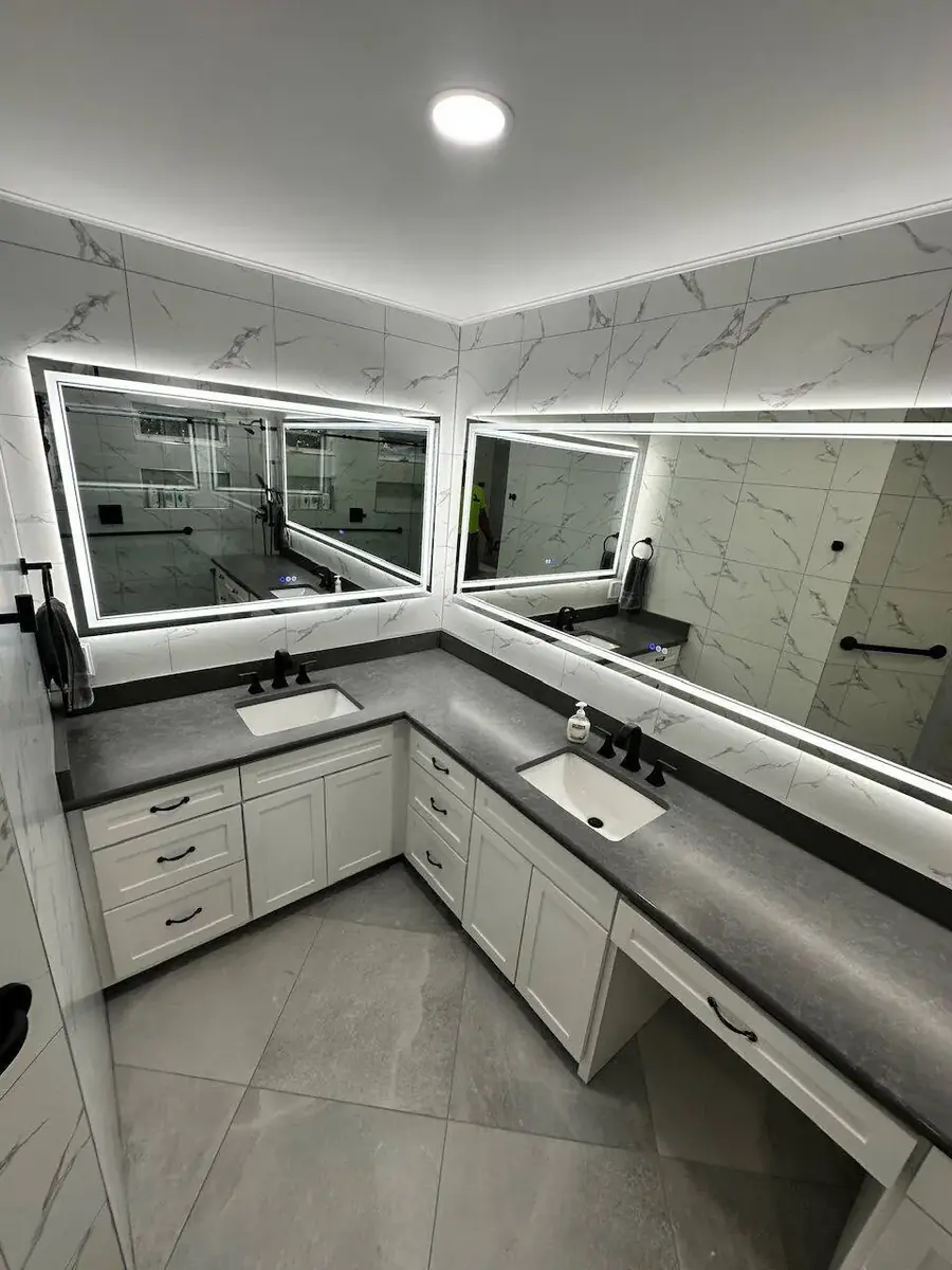 Completed master bathroom remodel in Metairie Louisiana with L-shaped dual-sink vanity, LED backlit mirrors, marble-look wall tile, gray stone-look countertops, and matte black fixtures by Cajun Home Services